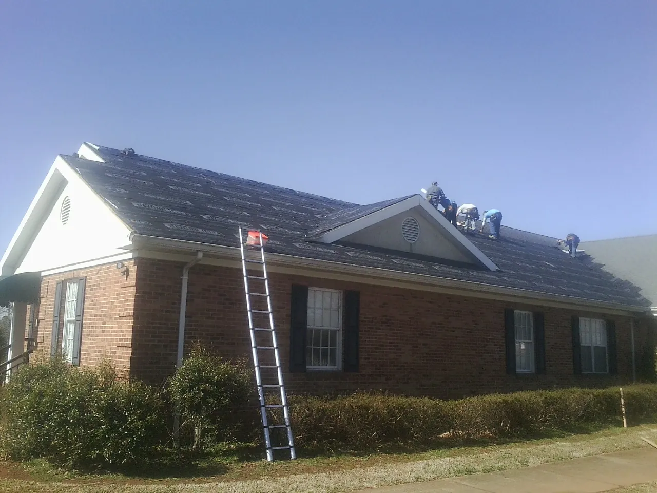 Crew tearing shingles off a brick ranch roof with a ladder set — Emergency Roof Repair work by our crew in Cabazon