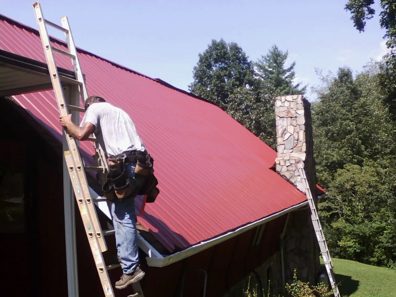 Roofer on a ladder beside a red metal roof and stone chimney — Soffit & Fascia Repair by Roofing Brothers in Cabazon